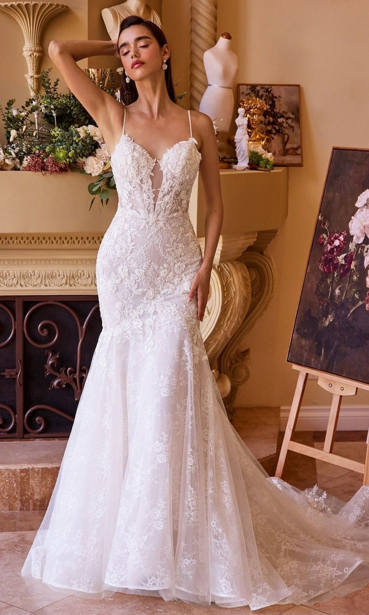 A woman poses indoors near a decorated mantel and floral painting, showcasing the delicate lace applique details of the Andrea and Leo WL051 fitted mermaid wedding gown with thin straps.